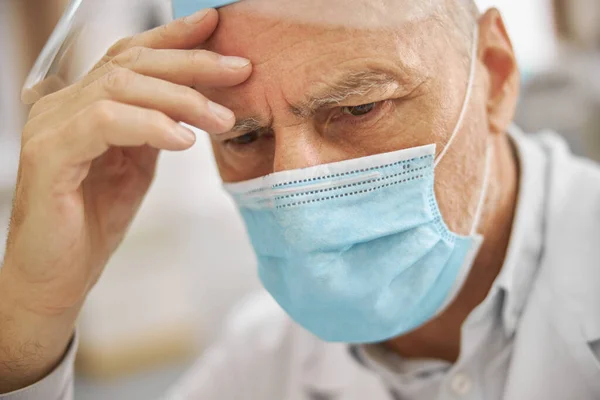 Ciudadano mayor usando una bata de laboratorio y una máscara que parece cansado — Foto de Stock