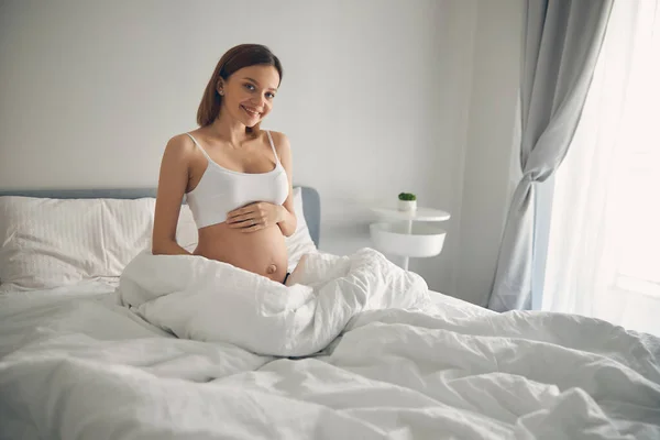 Smiling young lady caressing her belly in the white apartment — Stock Photo, Image