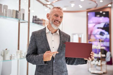 Dost canlısı yaşlı vatandaş dükkanda kırmızı bir kutu gösteriyor.