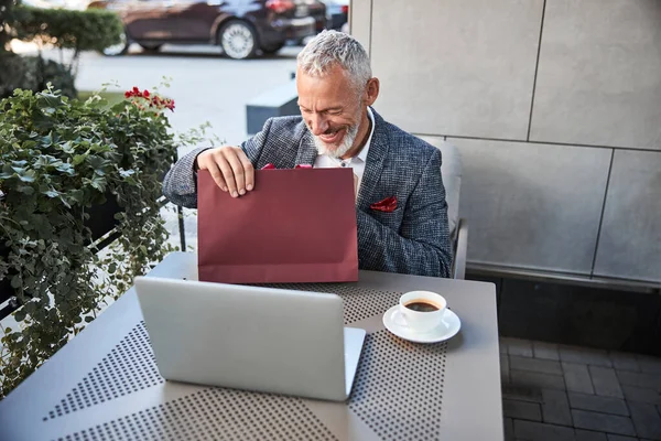 Joyful aging man checking the insides of a red gift bag — Stock Photo, Image