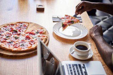 Afro-Amerikalı adam yemek yiyor ve pizzacıda dizüstü bilgisayar kullanıyor.