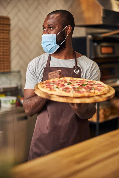 Uomo afro americano in maschera protettiva contenente pizza al salame piccante — Foto Stock