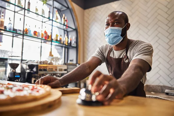 Male worker in medical mask ringing service bell in pizzeria — Stock Photo, Image