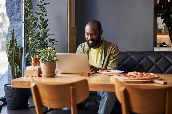 Alegre hombre afroamericano usando un portátil moderno en la cafetería — Foto de Stock