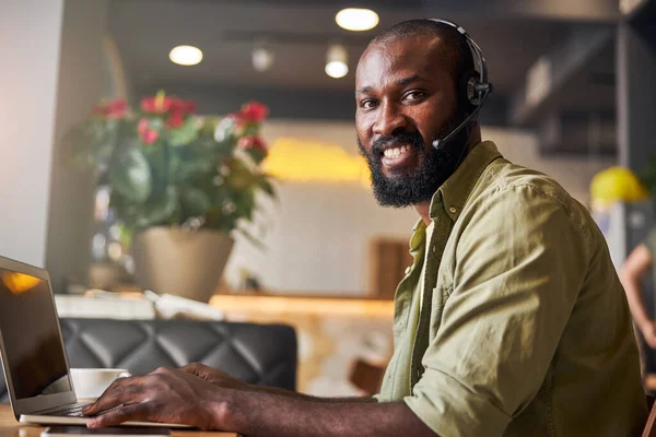 Alegre hombre afroamericano en auriculares usando el ordenador portátil en la cafetería —  Fotos de Stock