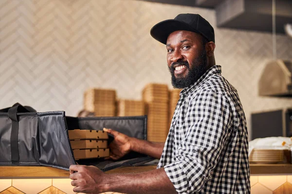 Homem afro-americano alegre colocando caixas de pizza no saco de entrega — Fotografia de Stock