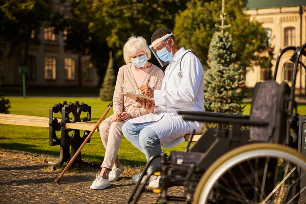 Cheerful medical specialist writing in notepad for his aging patient — Stock Photo, Image