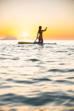 Calm young woman paddleboards on serene ocean waters during sunset. The tranquil scene is illuminated by golden hues, creating a peaceful atmosphere.