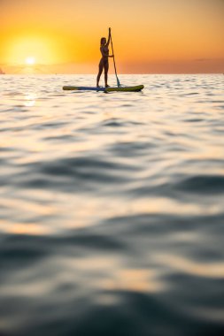 Young Caucasian woman stands on paddleboard in calm ocean at sunrise. Peaceful atmosphere captures tranquility and connection with nature.