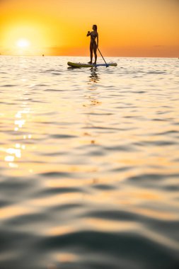 Young woman enjoys paddleboarding on calm ocean during sunset. The tranquil scene captures her serene expression as she gracefully navigates the shimmering water.