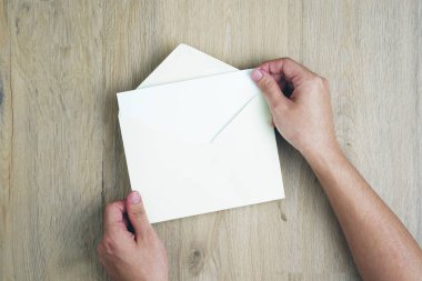 Bir adam el ele tutuşup ahşap masanın üstündeki zarfı açar..