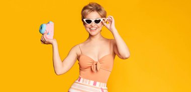 Yaz tatili. Güneş gözlüklü kadın gülümsüyor ve eski moda kamerayla fotoğraf çekiyor.