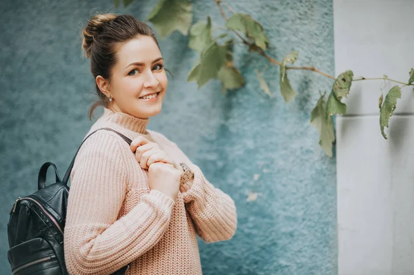 Outdoor portrait of young beautiful woman, wearing warm soft pullover, holding backpack, posing against blue wall