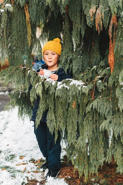 快乐的小男孩玩雪外面 穿着温暖的冬季夹克和黄色的帽子 孩子玩雪球 躲在树后 — 图库照片