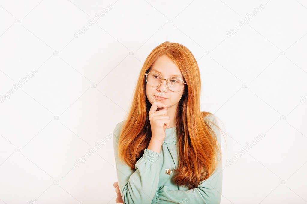 Studio shot of young preteen red-haired girl against white background, holding hand on the chin, looking far away