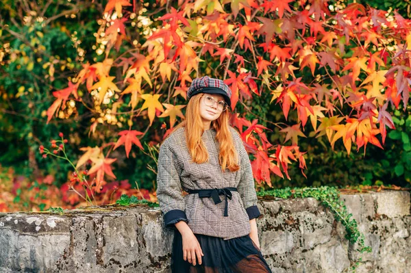 愛らしい小さな赤い髪の少女 オレンジ色の明るい木の横にポーズの屋外のポートレートの葉 チェックのジャケット 黒のスカートと帽子を身に着けています — ストック写真