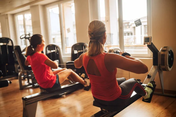 Back view of young fit women working out with rowing machine at gym