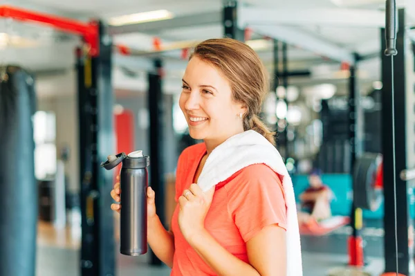 Happy and tired woman in sport gym, holding white towel and bottle of water