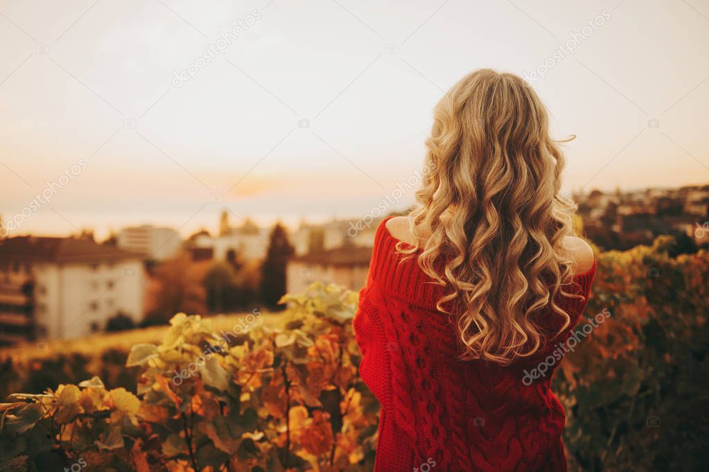 Outdoor portrait of woman with blond curly hair admiring sunset, wearing red pullover with open shoulders, back view. Image taken in Lavaux vineyards, Lausanne, Switzerland