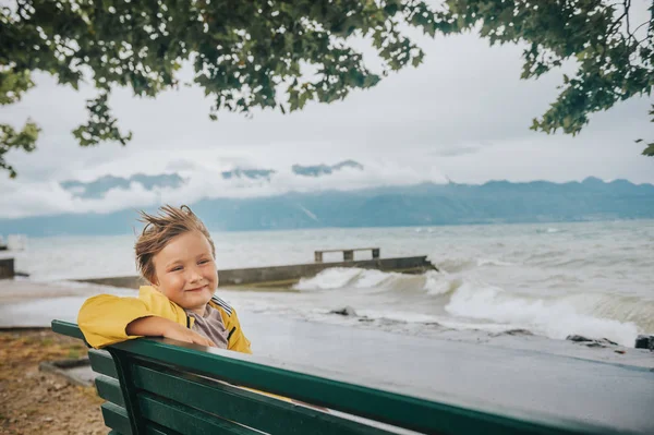 かわいい男の子非常に風の強い日は 湖のそばのベンチで休んで 波を見て 黄色の雨コートを着てします 湖ジュネーブ ローザンヌ スイス連邦共和国で撮影した画像 — ストック写真