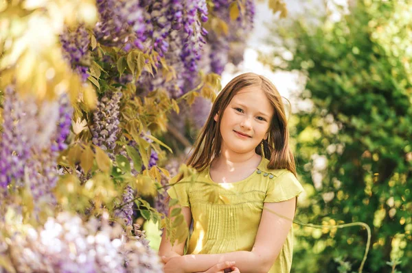 可爱的小女孩的 站在美丽的紫藤萝花 身穿绿色上衣的户外时尚肖像 — 图库照片