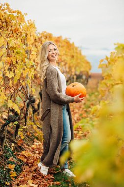 Sonbahar turuncu kabak holding, örme kahverengi longline hırka giymiş, altın üzüm bağları poz sarışın kıvırcık saçlı güzel kadın portresi