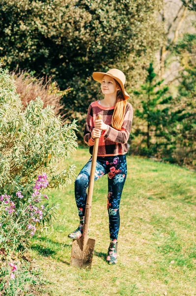 Outdoor portrait of young preteen kid girl helping in spring garden, holding a shovel, wearing a hat and fashionable rain boots