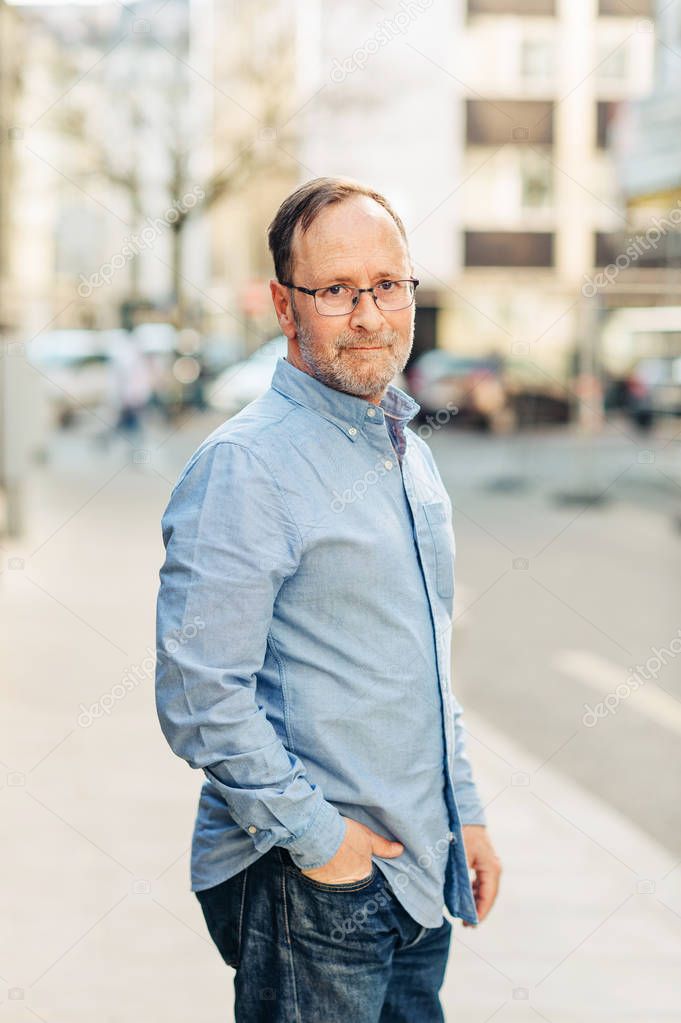 Outdoor portrait of handsome middle age man wearing long sleeve Oxford shirt, posing on city street, holding hand in jeans pocket