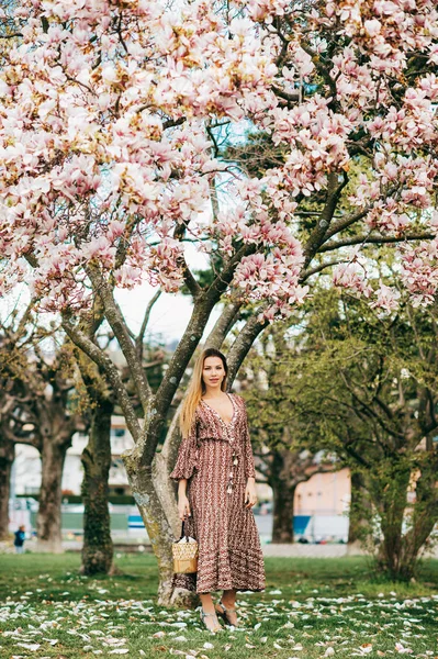 Spring portrait of fashionable young woman posing in spring park, wearing dress, holding lady bag