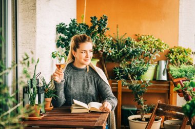 Rahat balkonda dinlenen, kitap okuyan, sıcak örme pullover giyen, bir kadeh şarap tutan genç güzel kadın 