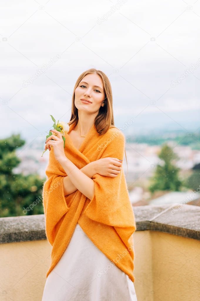 Romantic portrait of beautiful woman on the balcony under the summer rain, wearing bright ginger color knitted cape