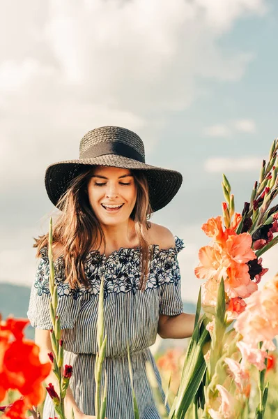 Muhteşem genç kadın bir alanda çiçek toplama, yazlık elbise, siyah hasır şapka giyiyor 