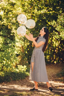 Esmer dalga saçlı güzel genç kadın, siyah beyaz elbise giyiyor, altın balonlar tutan