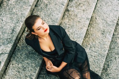 Fashion portrait of beautifl woman wearing black jacket and skirt, dark makeup with red lips, sitting on staire, top view clipart