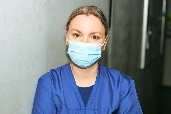Young female scrub nurse wear blue uniform and face mask, standing in hospital hallway, leaning on grey wall