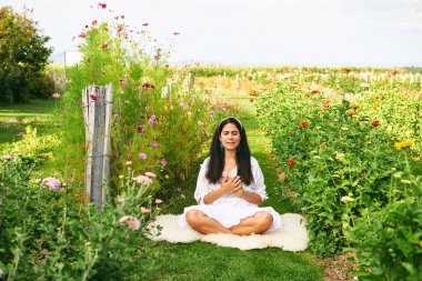 Orta yaşlı bir kadın yeşil bahçede meditasyon yapıyor, elleri kalbimde, gözleri kapalı.