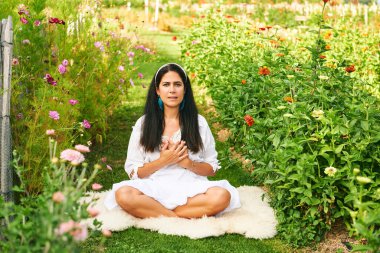 Orta yaşlı bir kadın yeşil bahçede meditasyon yapıyor, elini kalbe koyuyor, gözleri açık.