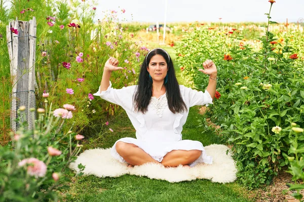 Orta yaşlı kadın yeşil bahçede meditasyon yapıyor, koyun derisinin üzerinde oturuyor.