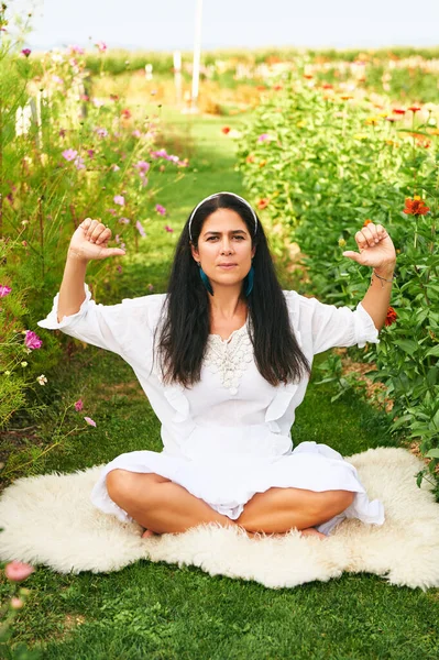 Orta yaşlı kadın yeşil bahçede meditasyon yapıyor, koyun derisinin üzerinde oturuyor.