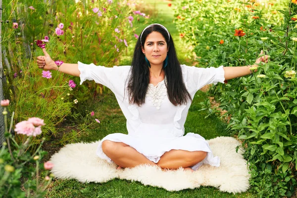 Orta yaşlı kadın yeşil bahçede meditasyon yapıyor, koyun derisinin üzerinde oturuyor.