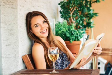 Genç mutlu kadın balkonda dinleniyor, kitap okuyor, duvara yaslanıyor, bir bardak şarap içiyor.