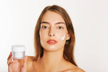 Yüzüne krem süren, beyaz arka plan, nemlendiricili kavanozu tutan genç ve güzel bir kadın resmi.