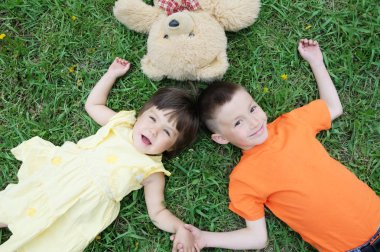 Üstten görünüm çocuklar eğleniyor Park'ta çim üzerinde yalan. Küçük kız ve erkek gülümseyen ile rahatlayın. Oyuncak ayı oyuncak birlikte