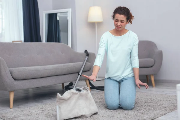 Dusty job. Young slender woman kneeling over open vacuum cleaner unwilling to touch dust and feeling disgusted by dirt