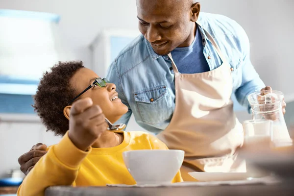 Angenehmer junger Vater plaudert mit Sohn, während er ihm Milch einschenkt — Stockfoto