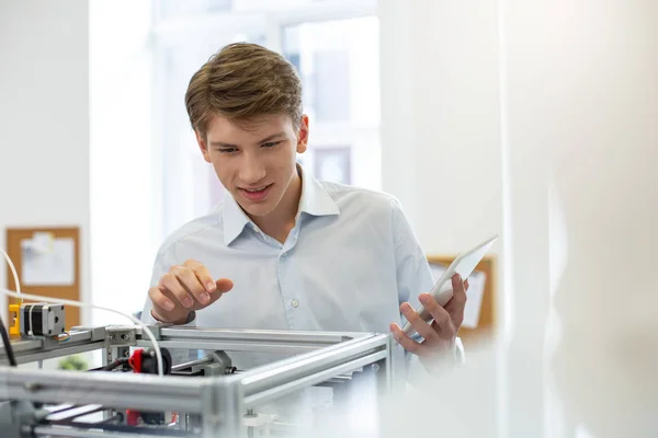 Молодой офисный работник улыбается и заглядывает внутрь 3D принтера — стоковое фото