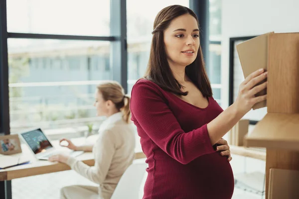 Attentive pregnant woman smiling and taking a book form the shelf — Stock Photo, Image