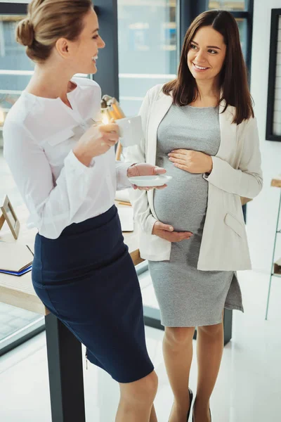 Cheerful secretary drinking coffee and her pregnant colleague smiling — Stock Photo, Image