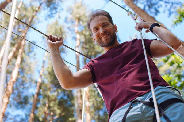 Cheerful young man smiling while looking down from rope trail — Stock Photo, Image
