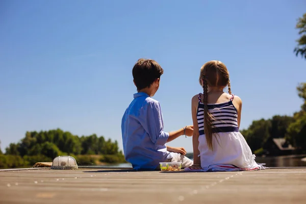Cute little siblings sitting on pier and having snacks — Stock Photo, Image
